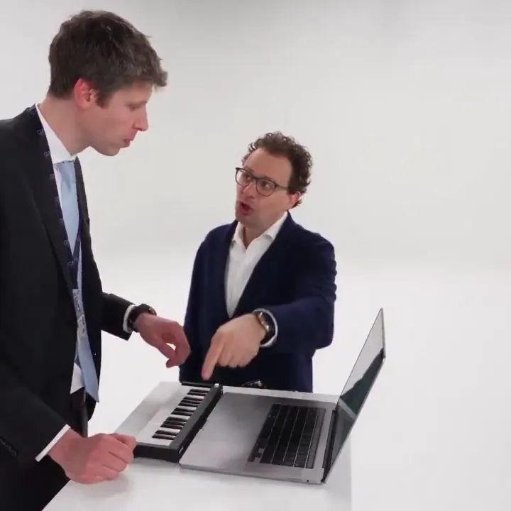 A man in a suit plays a mini keyboard next to a laptop while a friend in a navy cardigan points at the keys