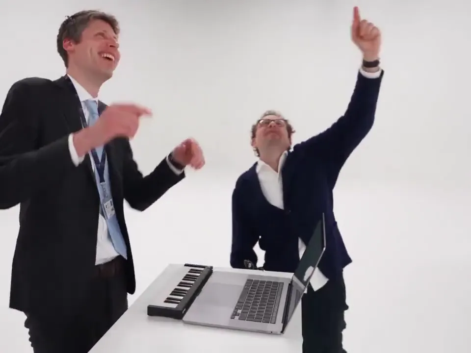 A man laughs and points at the ceiling while his friend throws an arm up in celebration at the keyboard