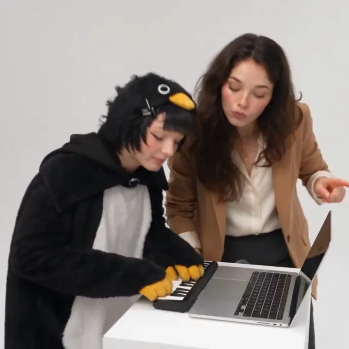 Two friends at a white desk with a laptop and mini keyboard in a white studio, one playing and one leaning in