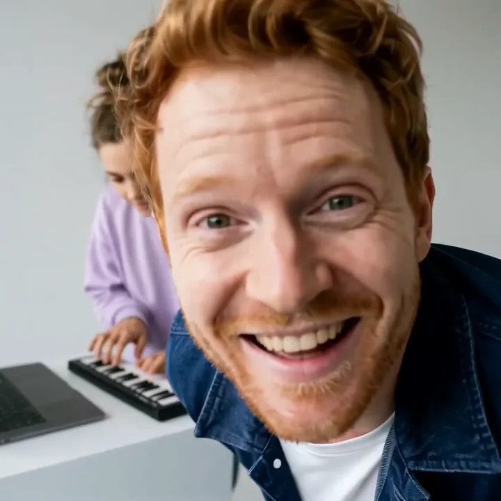 A red-haired man in a denim jacket grins into the lens so his face fills the frame, with his friend still at the keyboard behind him