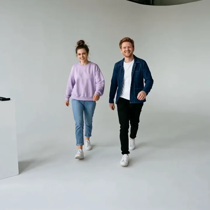 A woman in a lilac sweatshirt and a red-haired man in a denim jacket walk away from the desk across a white studio floor