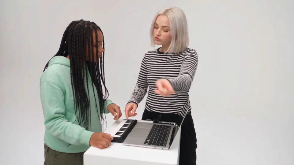 An Alors on Danse AI video: a woman with long braids in a mint hoodie plays a mini keyboard while her platinum-haired friend in a striped top points along, leans into the camera and winks, then both walk toward the camera in a white studio