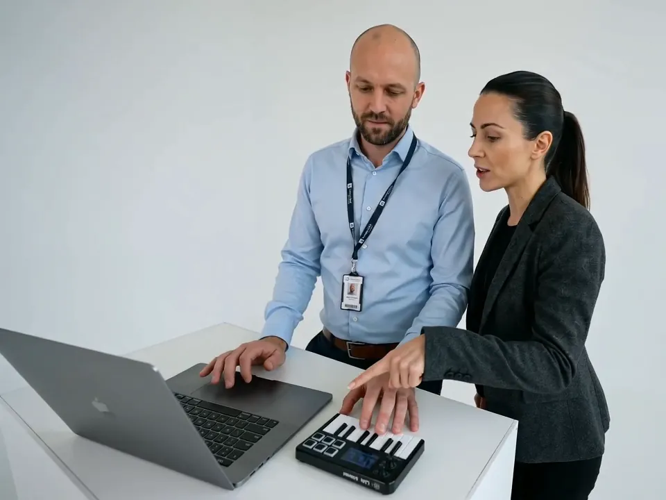 Two coworkers in office clothes, he with a lanyard badge, play the laptop and mini keyboard together