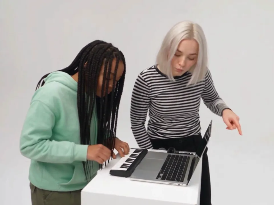 Two friends at the keyboard in the white studio: one with long braids in a mint hoodie plays while her friend in a striped top points